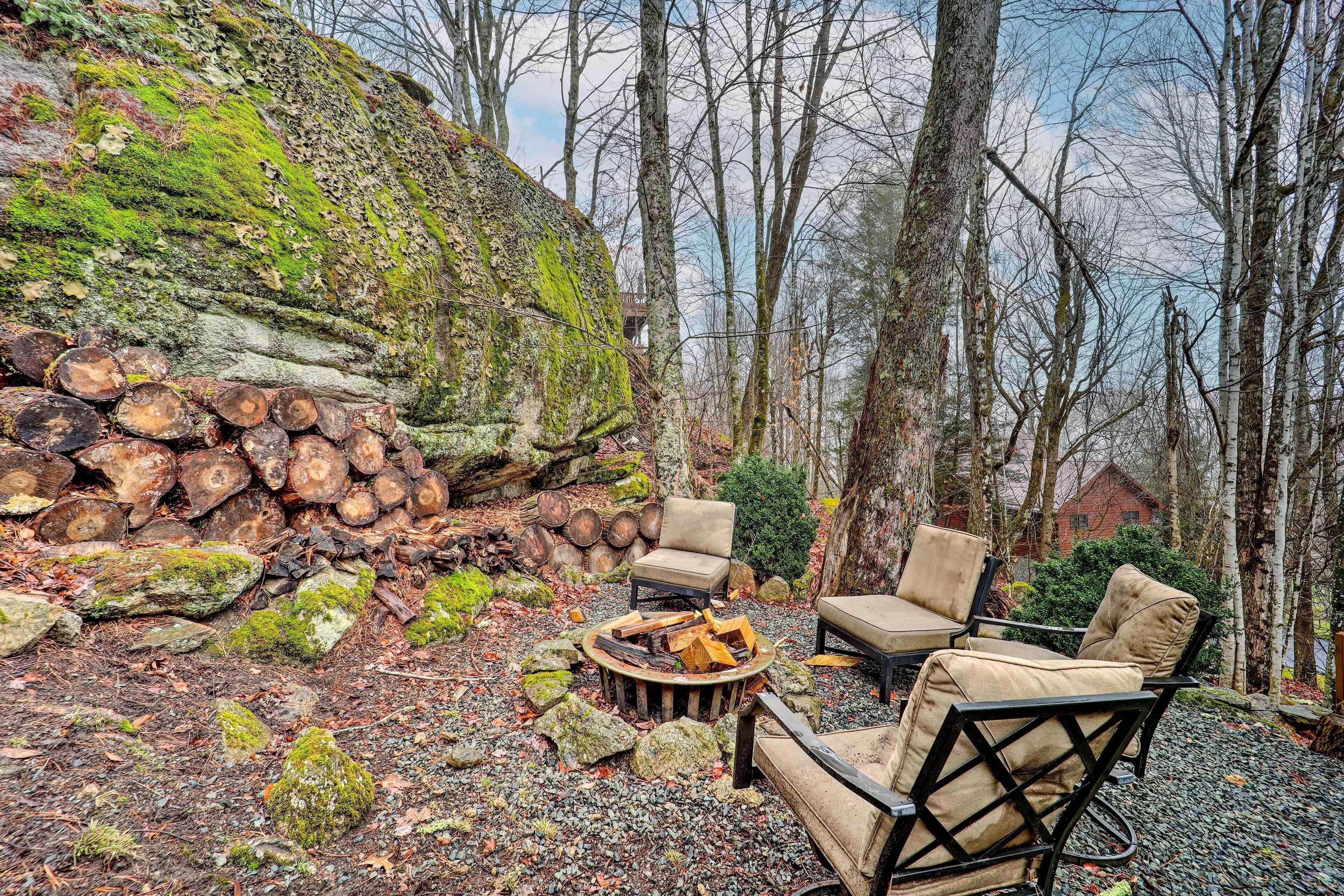 A rustic outdoor seating area with a fire pit, surrounded by logs and rocks, set against a backdrop of trees and a stone wall.