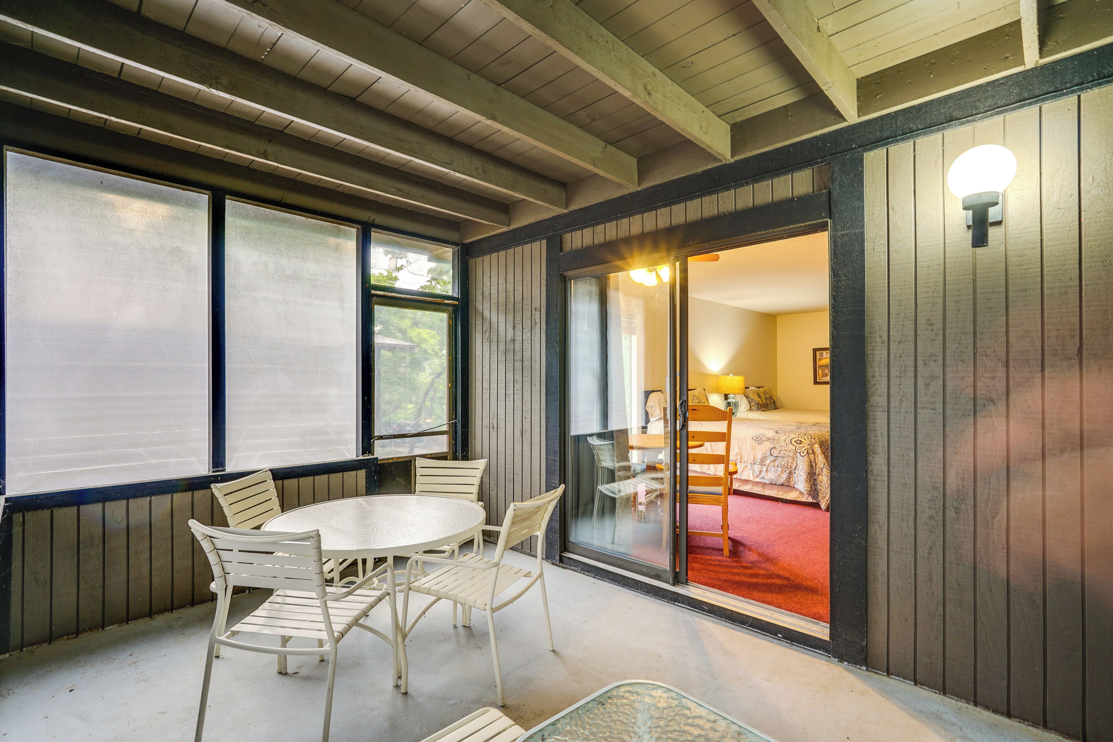 A room with a round table and chairs, a bed with a red bedspread, and a sliding glass door leading to another room.