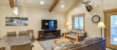 Una sala de estar con techo de madera, cabeza de venado montada, televisor, sofá y una mesa de comedor con sillas.