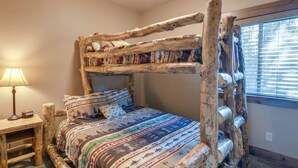 A rustic wooden bunk bed with a colorful quilt, a wooden nightstand with a lamp, and a window with blinds. - Outdoor Adventure Hub ~ 20 Miles to Yellowstone! (Island Park)