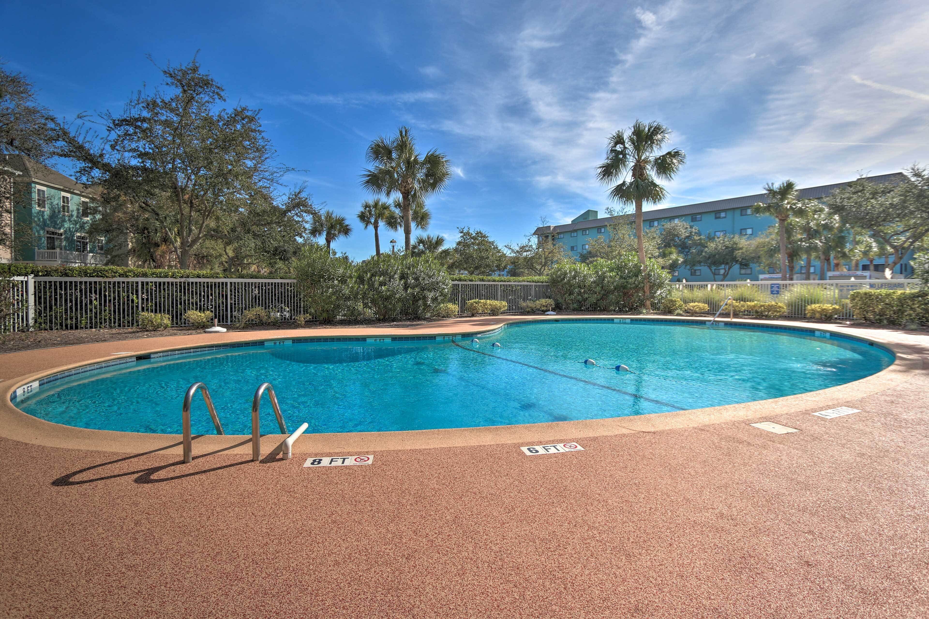 A swimming pool with a red deck, surrounded by palm trees and a building in the background.