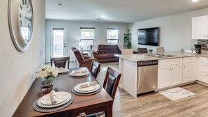 Eine moderne Küche mit integriertem Geschirrspüler, ein Essbereich mit Holztisch und -stühlen sowie ein Wohnzimmer mit braunem Sofa und Flachbildfernseher.