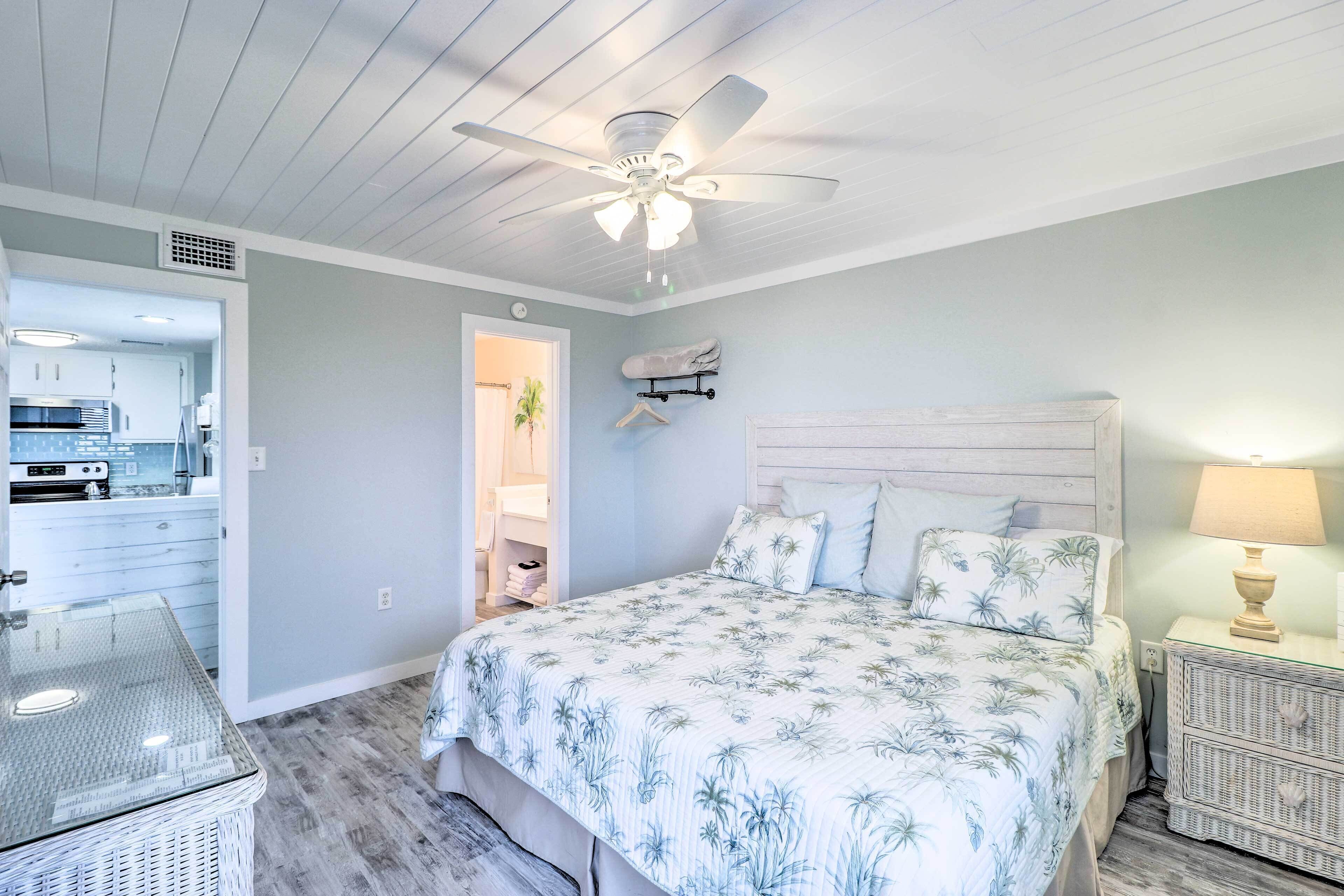A bedroom with a bed, a nightstand, a ceiling fan, and a view into the kitchen.