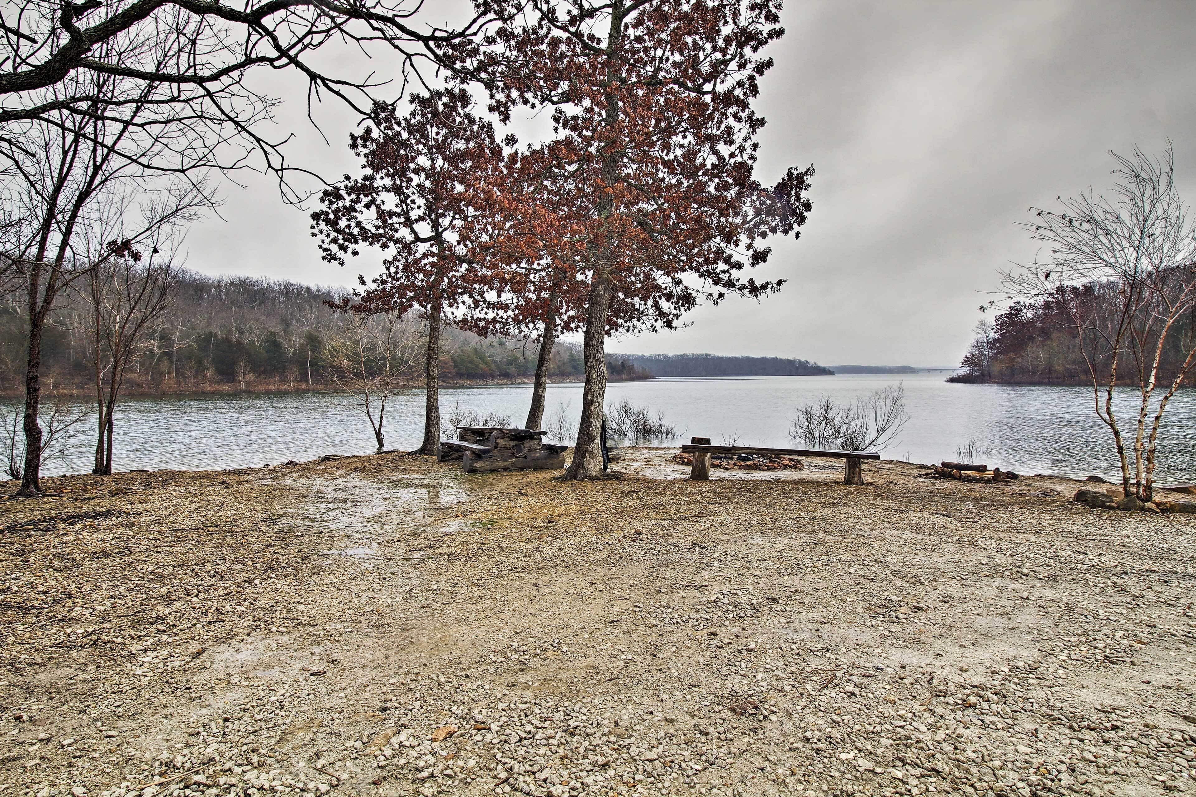 A lakeside park with benches and picnic tables, surrounded by trees and a view of the lake.