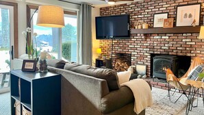A cozy living room with a brick fireplace, a flat-screen TV mounted above it, a comfortable sofa, a coffee table, and a chair. - Glen Condo w/ Mountain Views: Walk to Story Land (Glen)