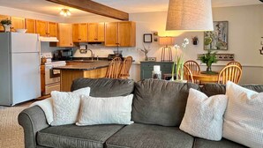 A cozy living room with a grey sofa, wooden dining table, and a kitchen area with wooden cabinets and a microwave. - Glen Condo w/ Mountain Views: Walk to Story Land (Glen)