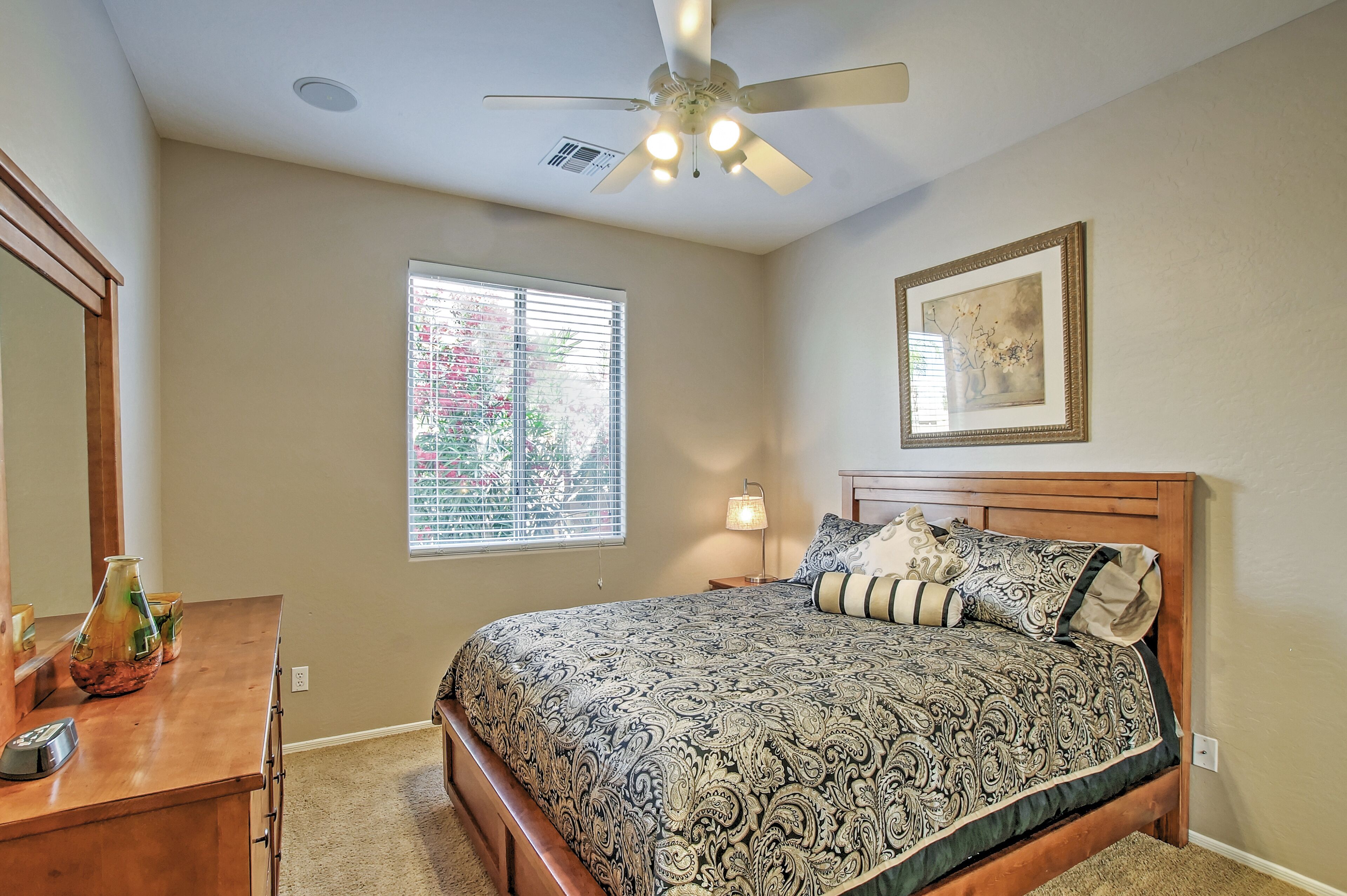 A bedroom with a bed, a ceiling fan, a window with blinds, a bedside lamp, and a framed picture on the wall.