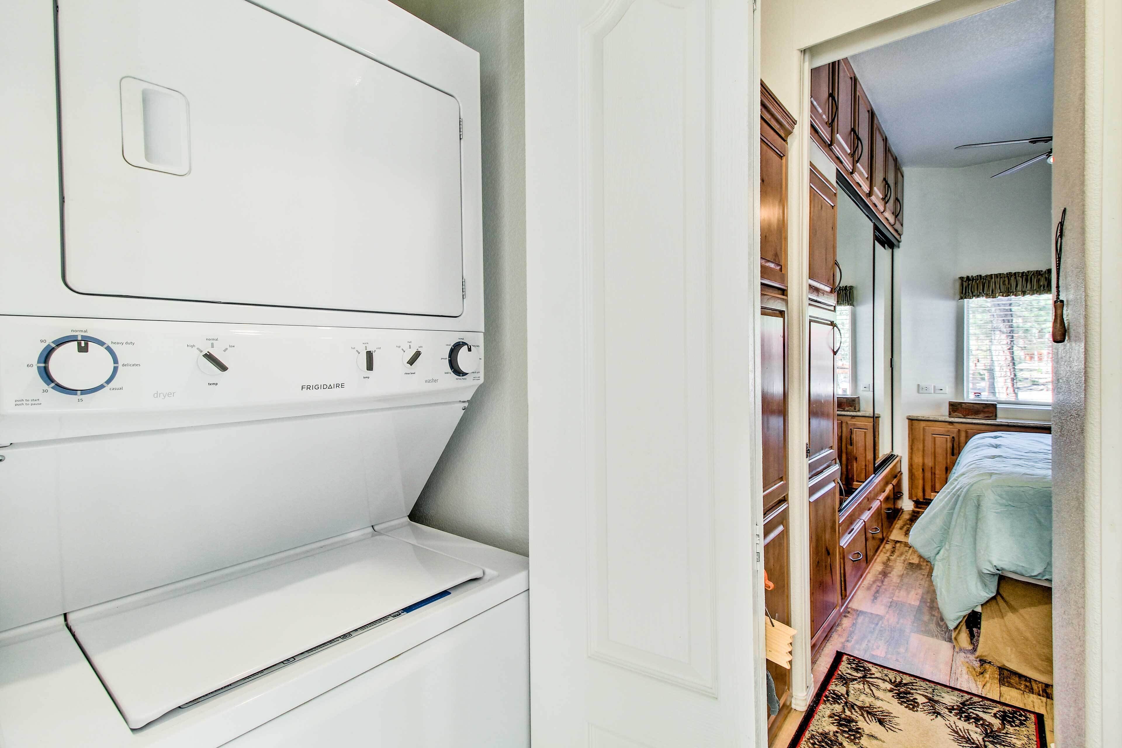 A laundry area with a white dryer and a washing machine. There is a wooden cabinet and a bed with a green blanket.