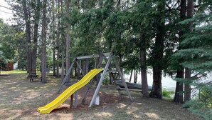 A wooden swing set with a yellow slide in a wooded park area. - Pine Ridge Resort Cabin on Bass Lake w/ Dock! (Interlochen)