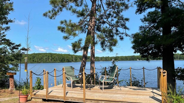 A wooden deck with chairs and a table, surrounded by a rope barrier, overlooking a lake. - Pine Ridge Resort Cabin on Bass Lake w/ Dock! (Interlochen)
