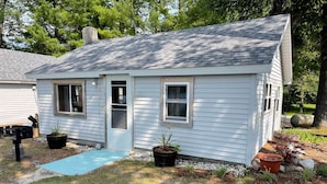 A small, single-story house with a sloped roof, a front door, and two windows. There is a blue mat leading to the door, and several potted plants are placed around the front yard. - Pine Ridge Resort Cabin on Bass Lake w/ Dock! (Interlochen)