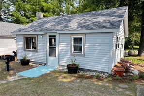 A small, single-story house with a sloped roof, a front door, and two windows. There is a blue mat in front of the door, and potted plants are placed near the entrance. - Pine Ridge Resort Cabin on Bass Lake w/ Dock! (Interlochen)