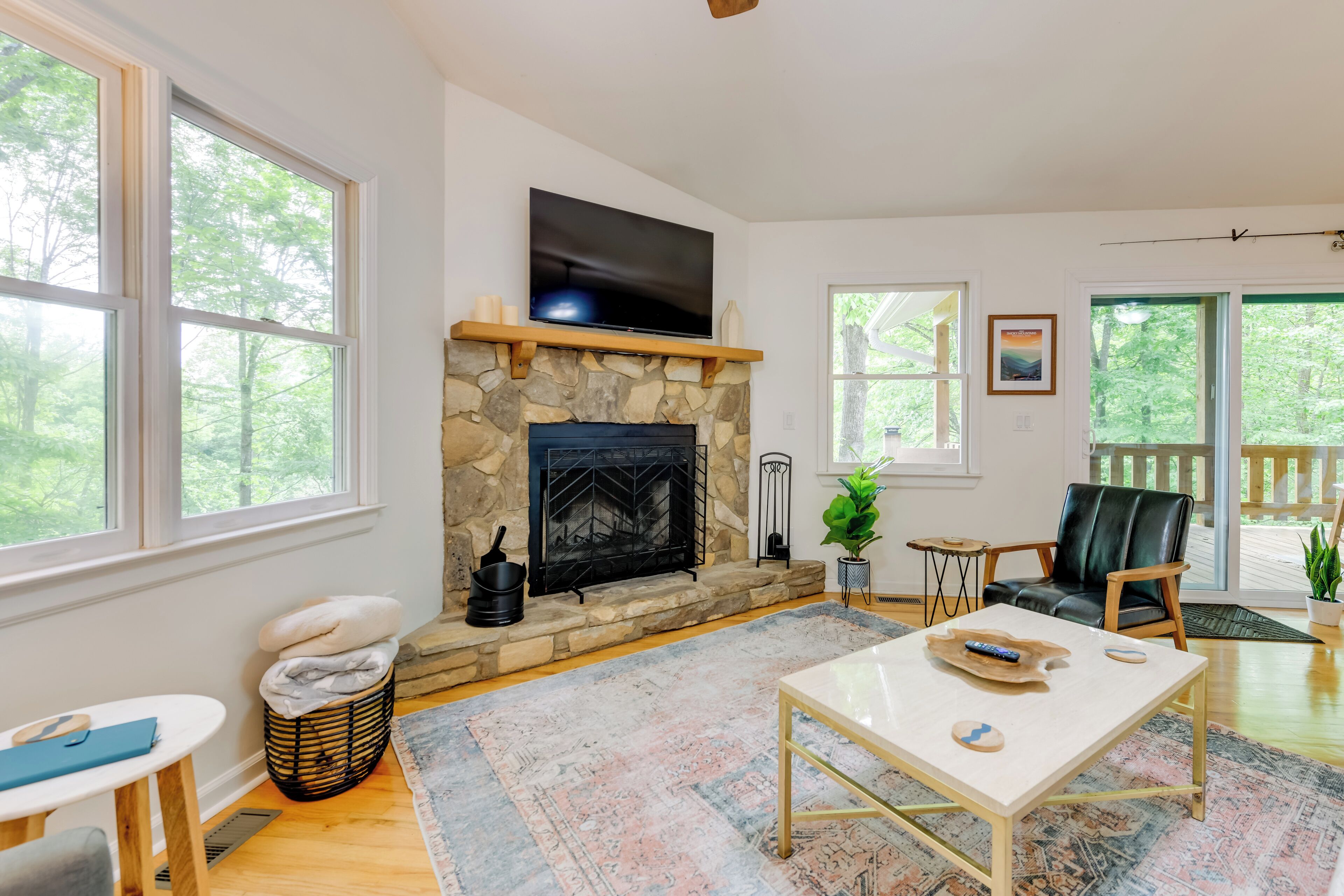 A living room with a stone fireplace, a flat-screen TV, a comfortable seating area, and large windows offering a view of the outdoors.