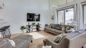 A modern living room with a large TV, comfortable seating, a small bar area, and a view through large windows. - Gorgeous Lakefront Townhome w/ Pool Access & Views (Montgomery)
