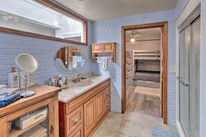 A bathroom with a wooden vanity, a large mirror, and a bunk bed visible through an open door.