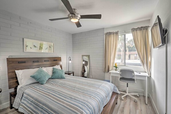Une chambre à coucher comprenant un lit, un bureau, une chaise, un ventilateur de plafond et une fenêtre avec des rideaux.