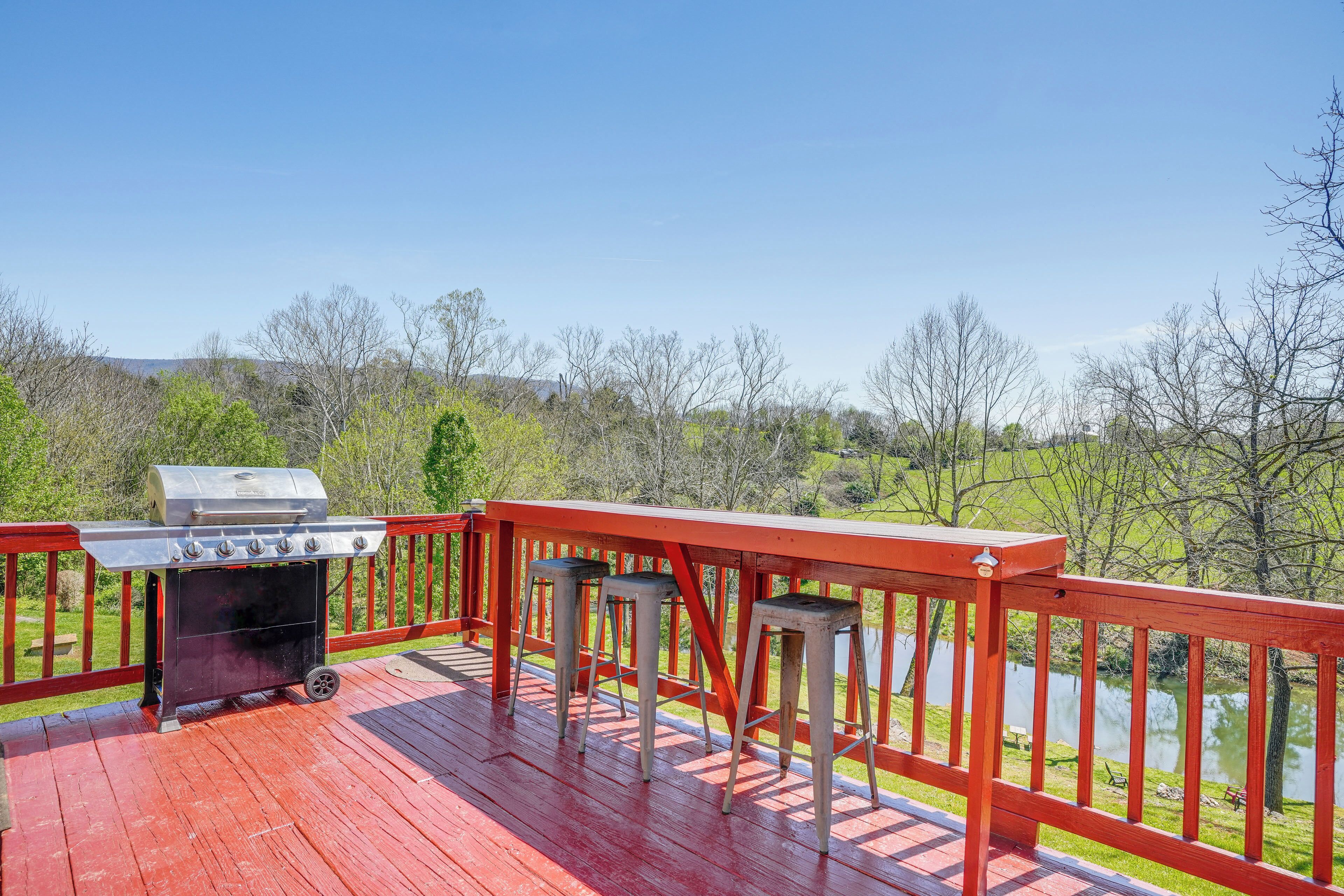 Une terrasse rouge avec un barbecue et des tabourets, d’où l’on a vue sur un lac et des arbres.