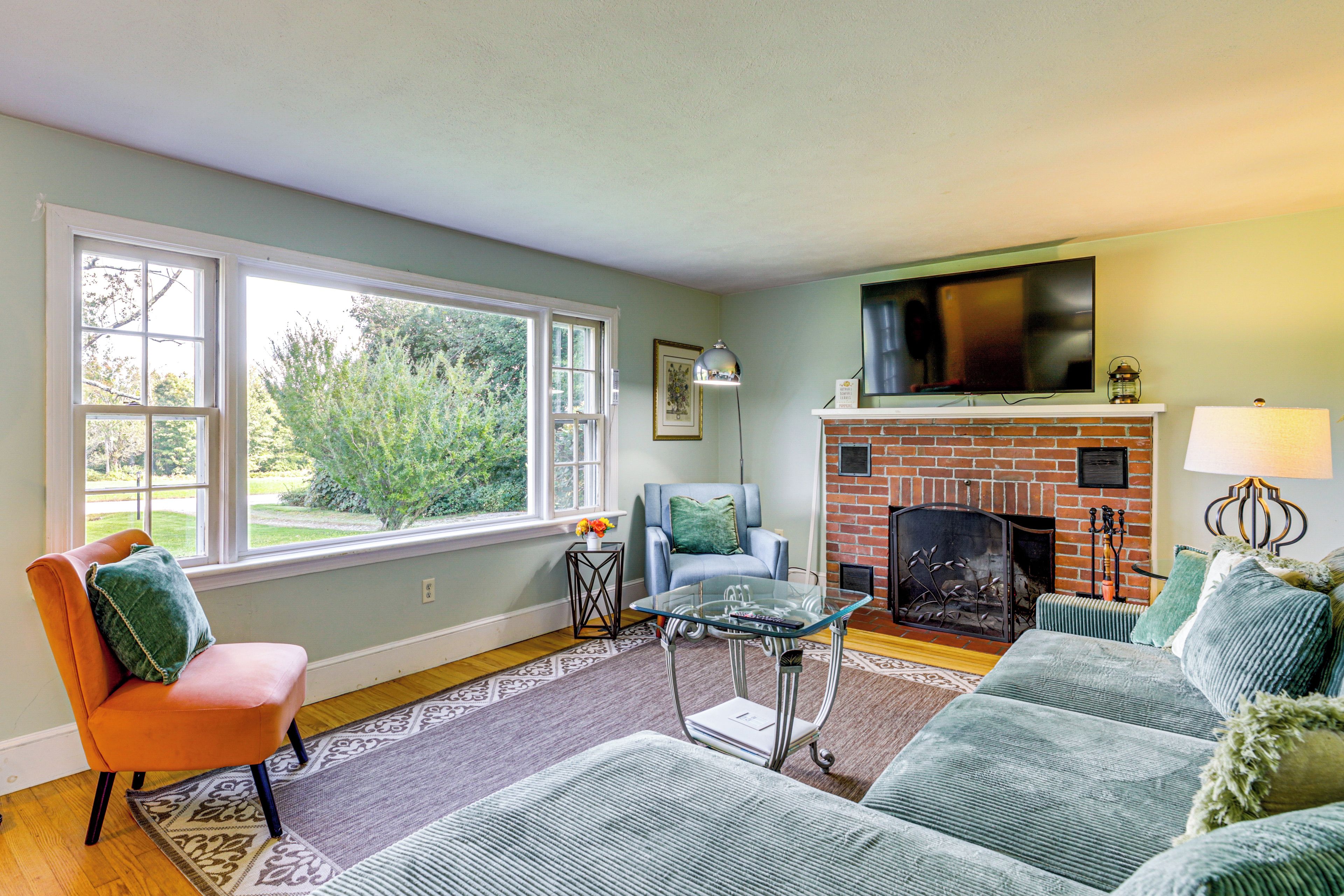 A living room with a fireplace, a television mounted above it, and a large window providing a view of the outdoors.