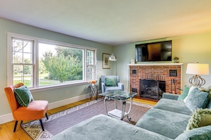 A living room with a fireplace, a television mounted above it, and a large window providing a view of the outdoors. - Hampton 'quiet Corner Cottage' w/ Fireplace & Yard (Hampton)