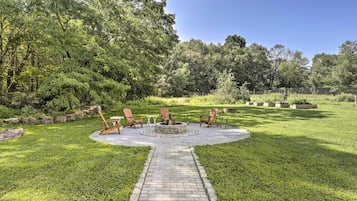 A circular outdoor seating area with a fire pit, surrounded by a brick pathway, and a grassy lawn with trees in the background.