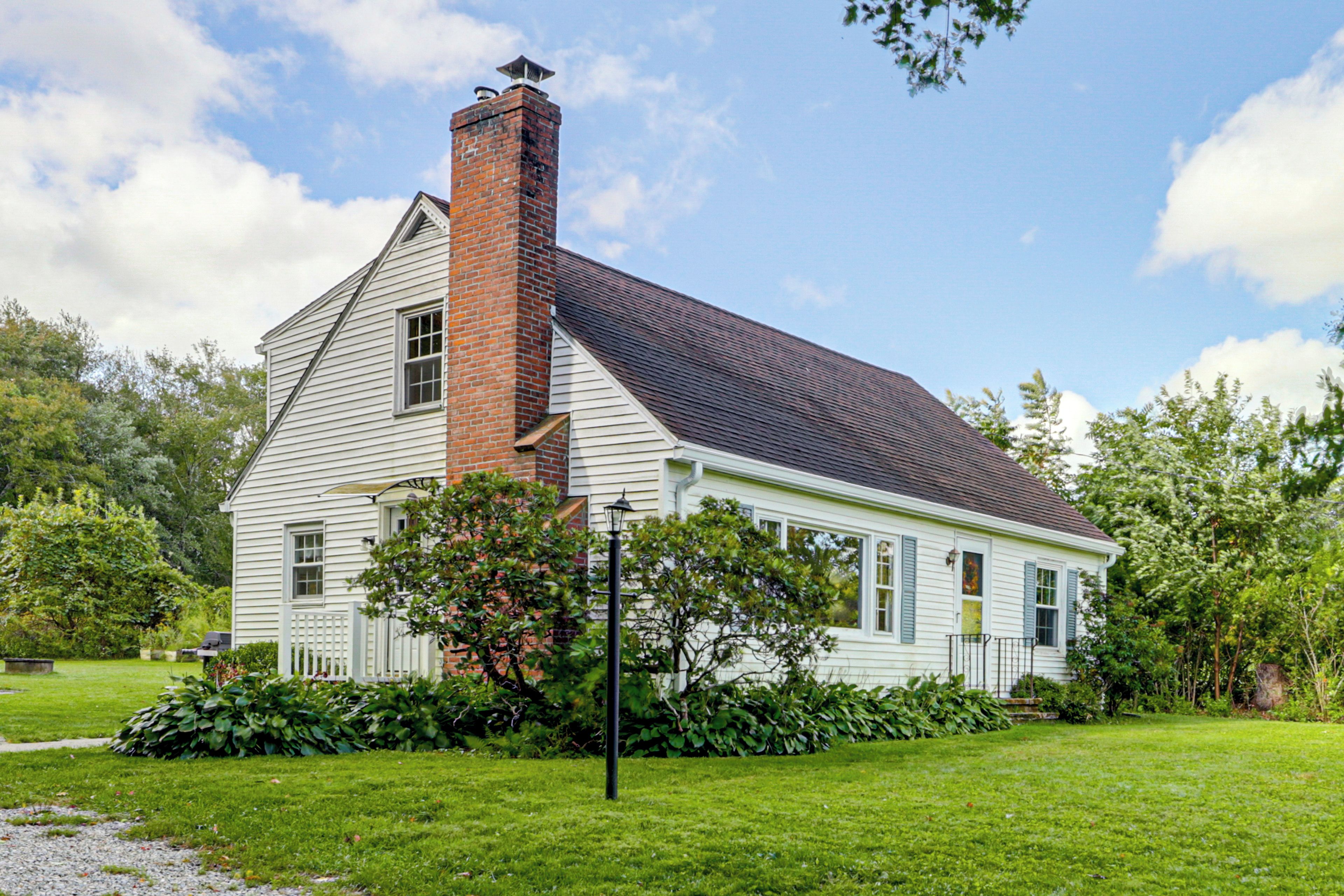 A single-story house with a brick chimney, white siding, and a dark roof.