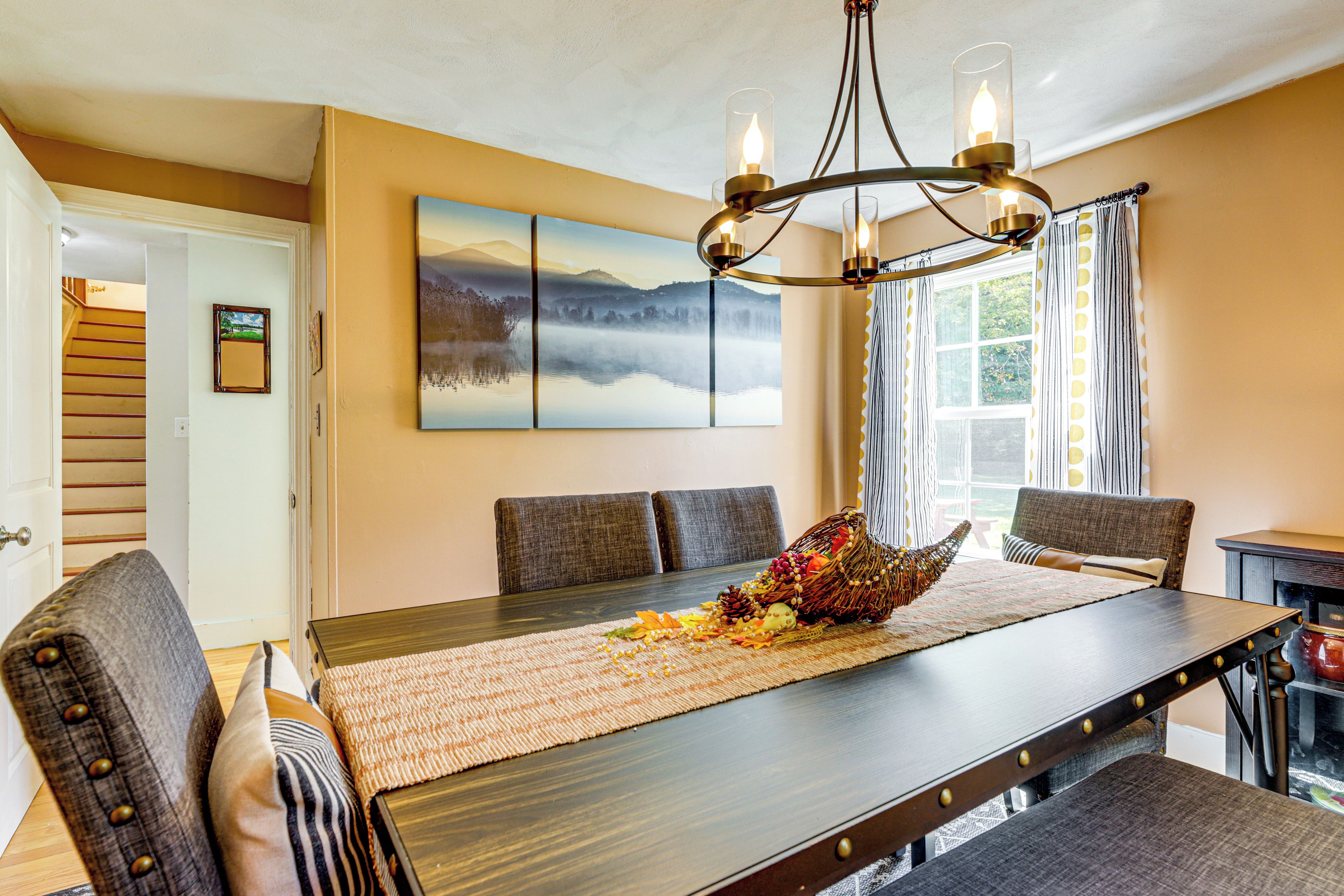 A dining area with a wooden table, cushioned chairs, a chandelier, and a large landscape painting on the wall.