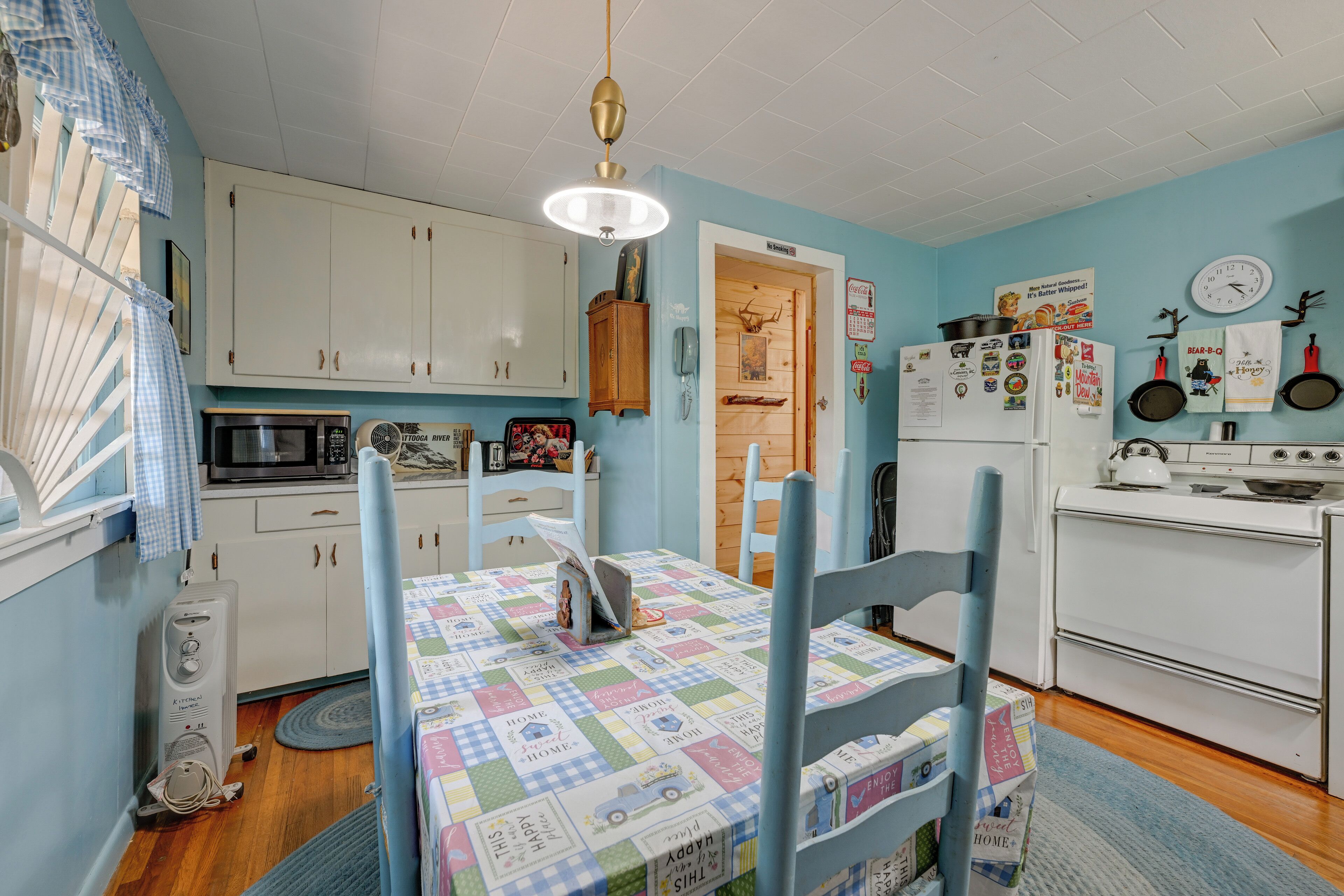 A kitchen with white cabinets, a microwave, a refrigerator, and a stove. There is a table with a colorful tablecloth and blue chairs.