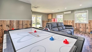 A pool table with shuffleboard pucks, a leather couch, and wooden paneling in a room with large windows. - Walk to Lake George: Family Escape w/ Game Room! (Lake George)