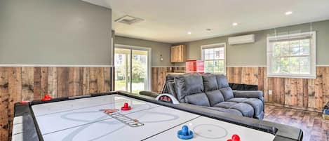 A pool table with shuffleboard pucks, a leather couch, and wooden paneling in a room with large windows.