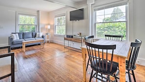 A spacious living room with hardwood floors, a gray sofa, a wooden dining table with chairs, and large windows offering a view of greenery. - Walk to Lake George: Family Escape w/ Game Room! (Lake George)