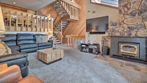 A spacious living area with a black leather sofa, a wooden coffee table, a stone fireplace, a TV mounted on the wall, and a wooden staircase leading to an upper level. - The Mammoth Lodge by Hiking, Lakes, Skiing & More! (Mammoth Lakes)