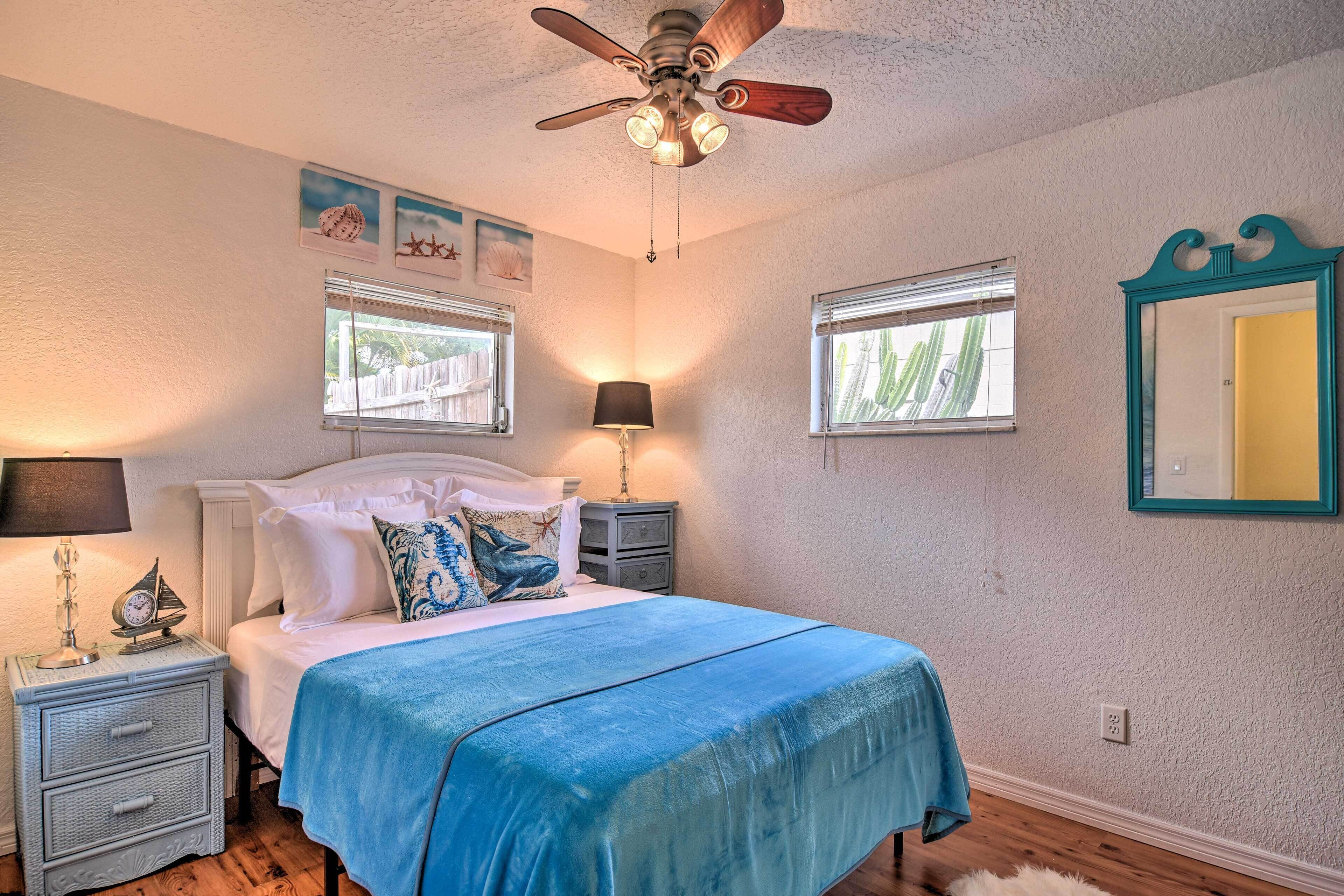 A bedroom with a bed, nightstands, a ceiling fan, two windows, and wall art.