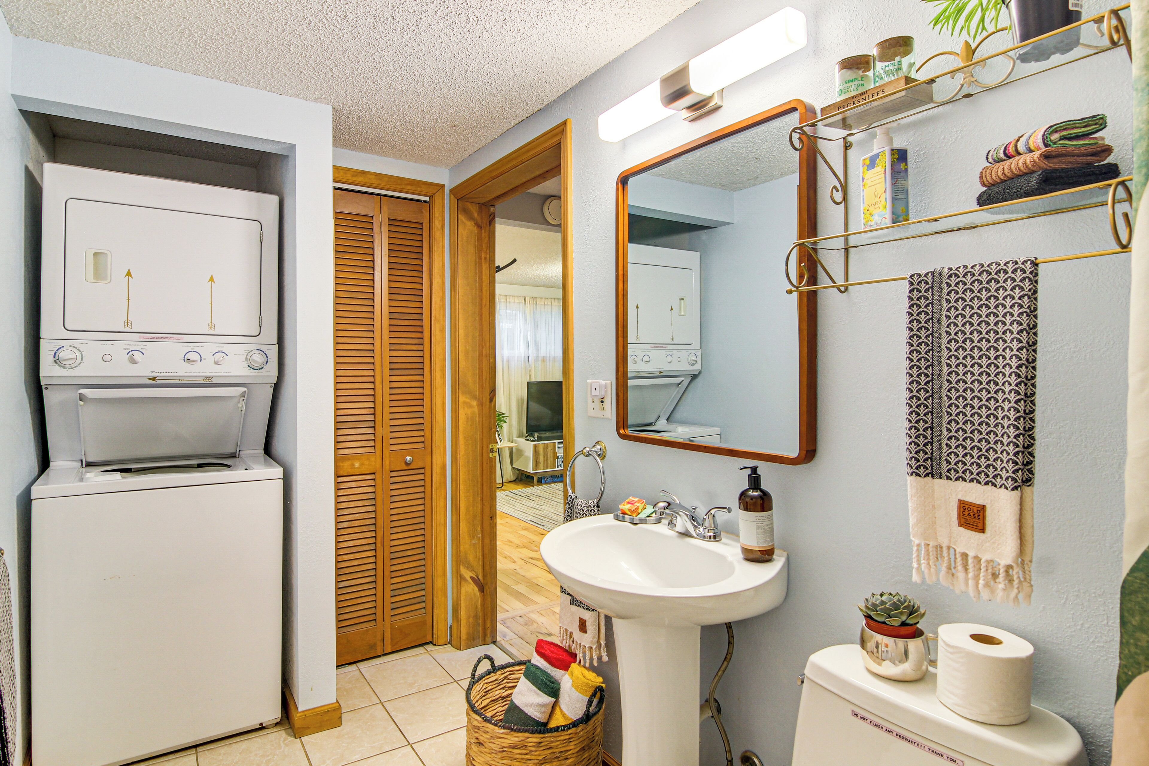 Une petite salle de bain comprenant un lave-linge blanc, un lavabo, un miroir et une étagère avec des serviettes et des articles de toilette.