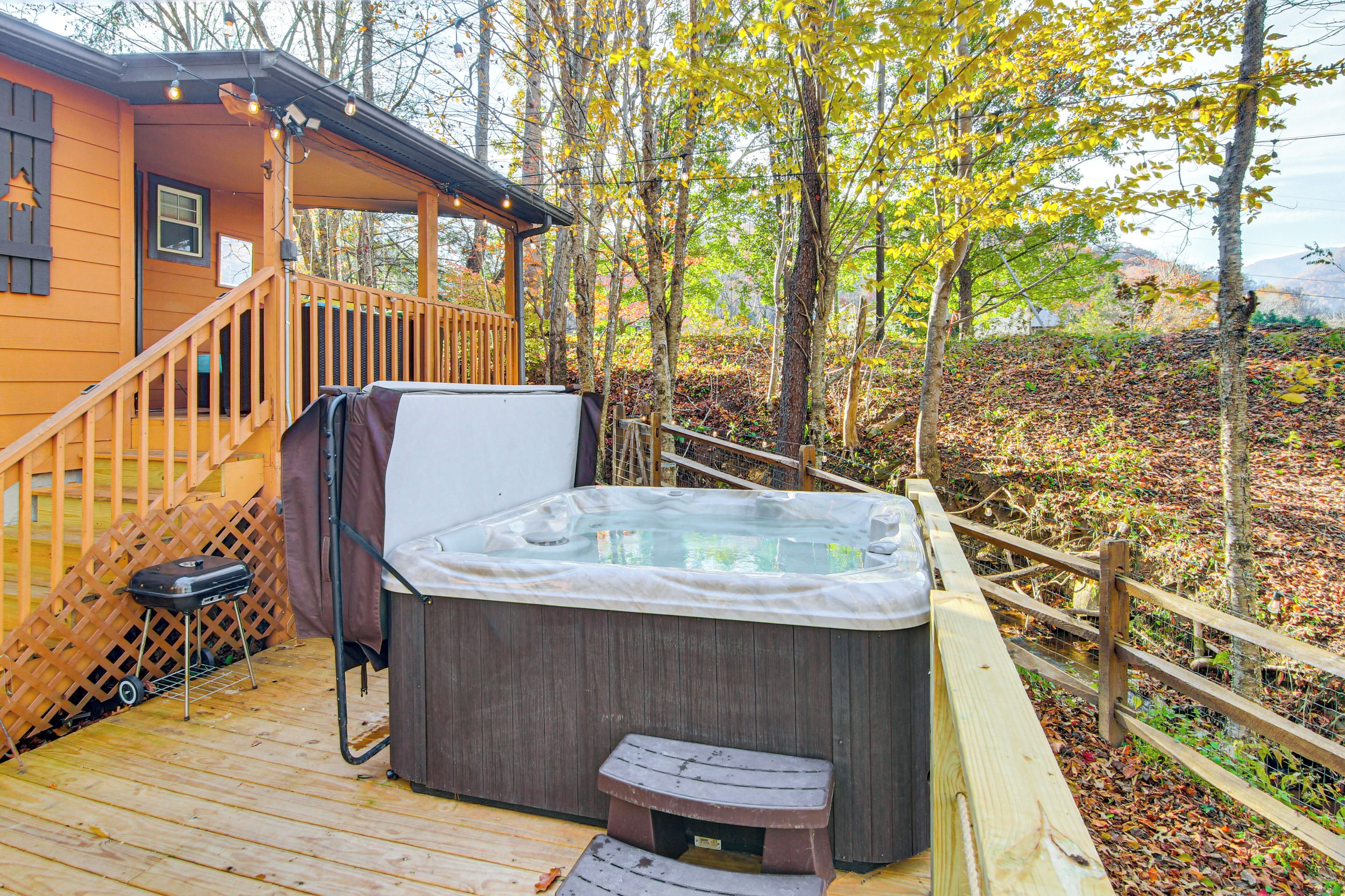Une terrasse en bois avec un jacuzzi, un espace salon couvert et une vue sur une zone boisée.
