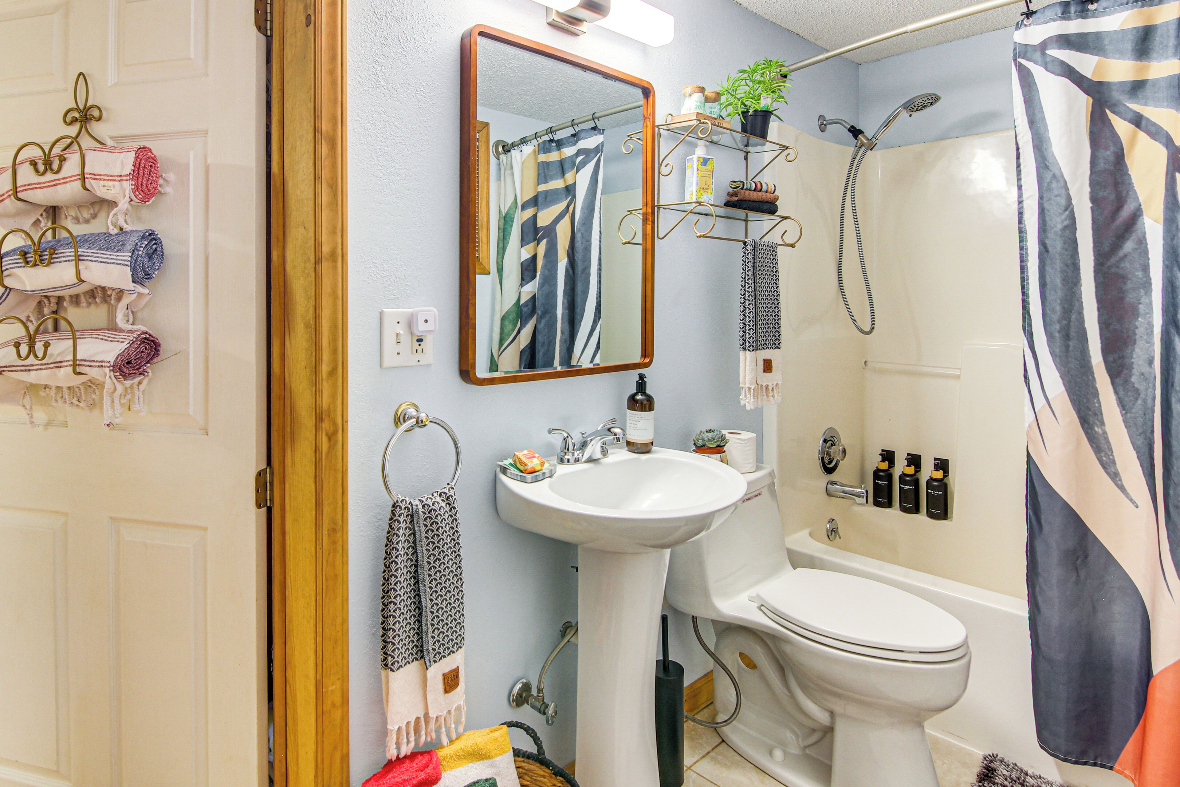 A bathroom with a sink, toilet, and shower. There are towels hanging on a rack, a mirror, and a shelf with toiletries.