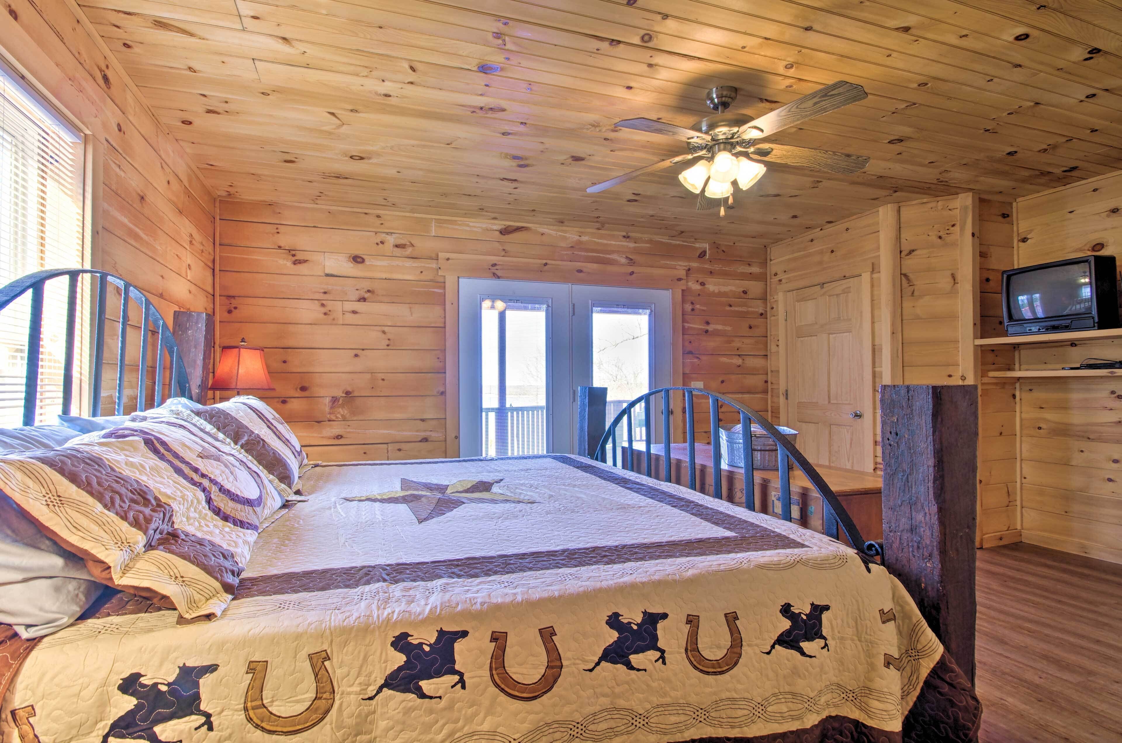 A wooden cabin room with a bed, a ceiling fan, a television, and a window.