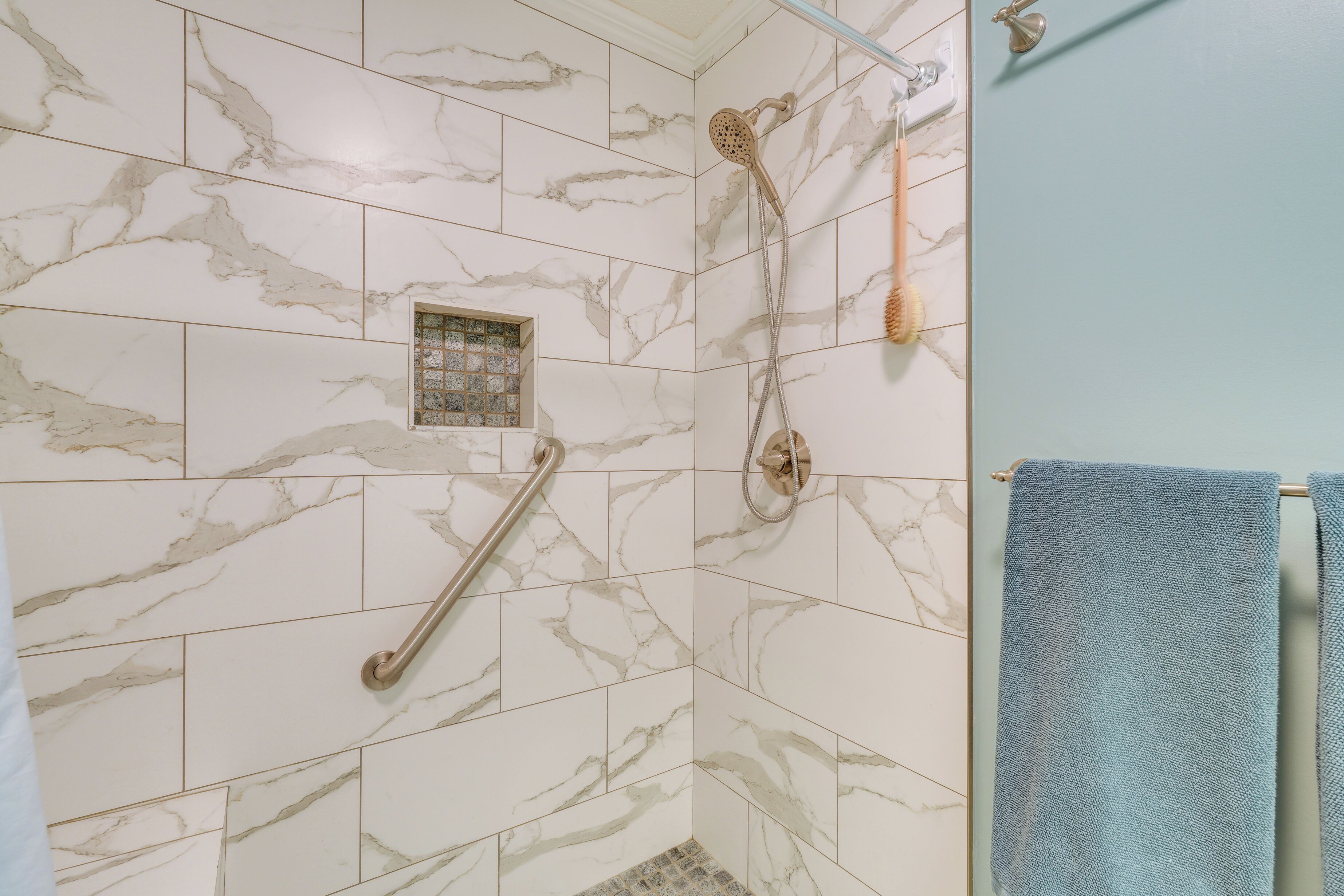 Une salle de bain carrelée de marbre, une douche avec un pommeau de douche amovible, une barre d’appui et un porte-serviettes avec une serviette bleue.