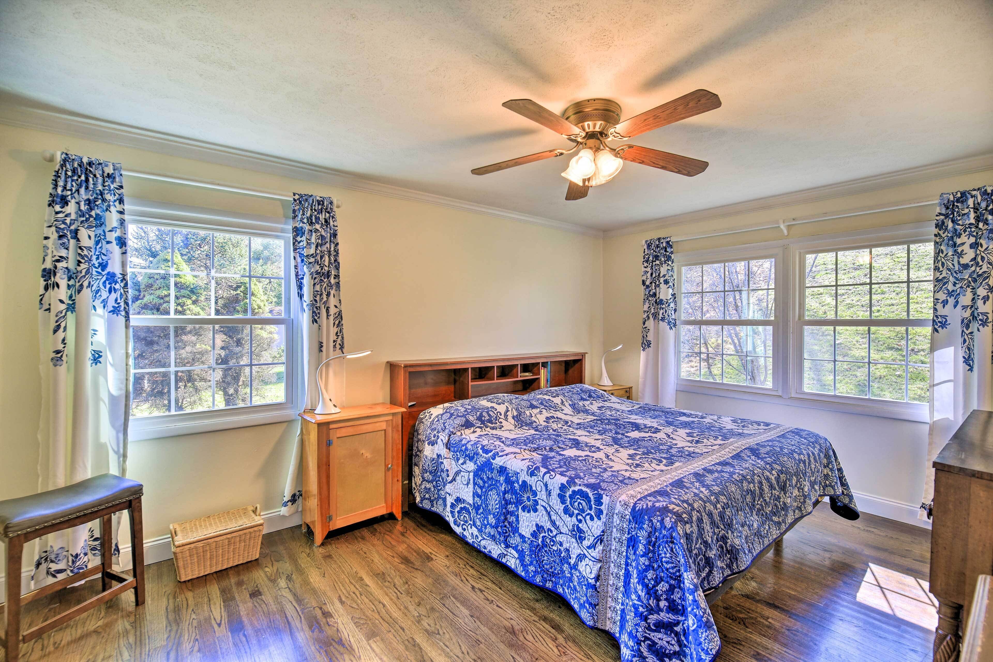A bedroom with a bed, wooden headboard, two windows with curtains, a ceiling fan, and a nightstand.