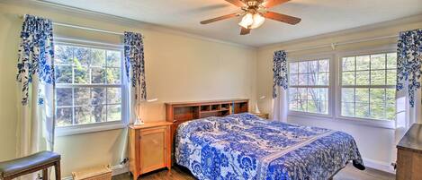 A bedroom with a bed, wooden headboard, two windows with curtains, a ceiling fan, and a nightstand.