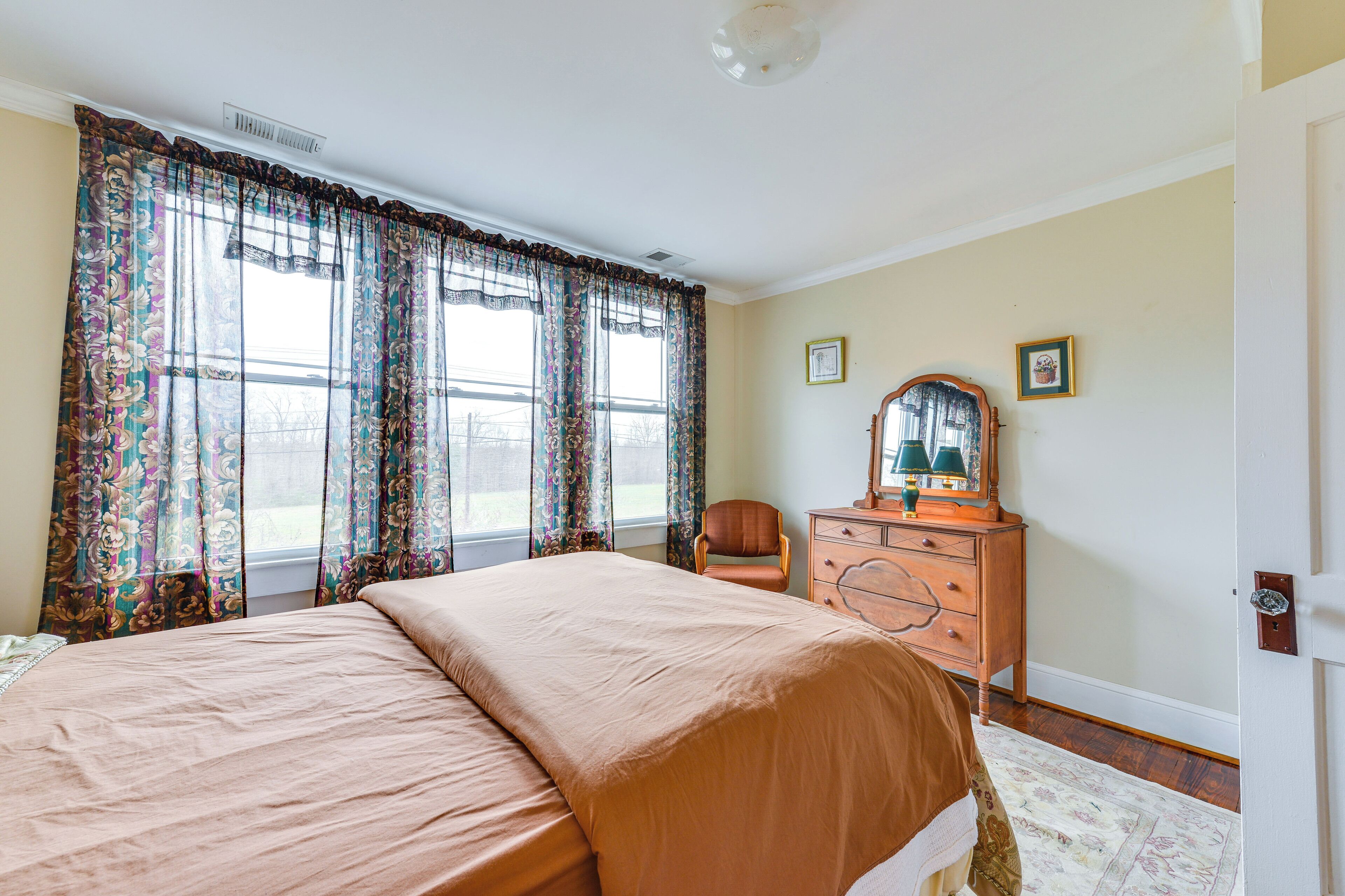 A bedroom with a large bed, a dresser, a chair, and a window with curtains.