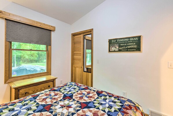 A bedroom with a large window, a wooden dresser, a quilted bedspread, and a wall sign advertising fishing gear. - Charming Thompsonville Home w/ On-site River! (Thompsonville)
