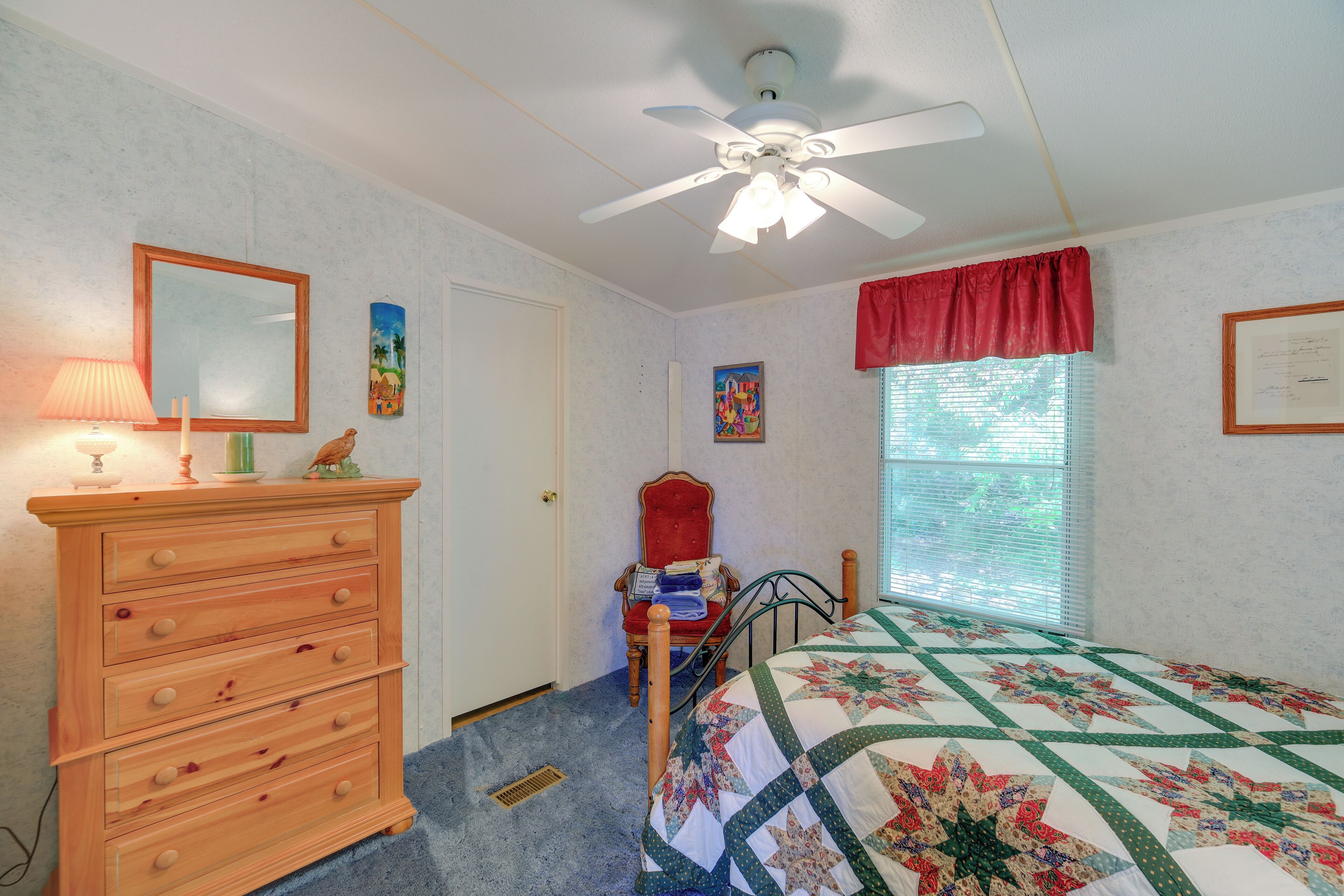 A bedroom with a wooden dresser, a bed with a quilt, a ceiling fan, a window with blinds, and a chair.