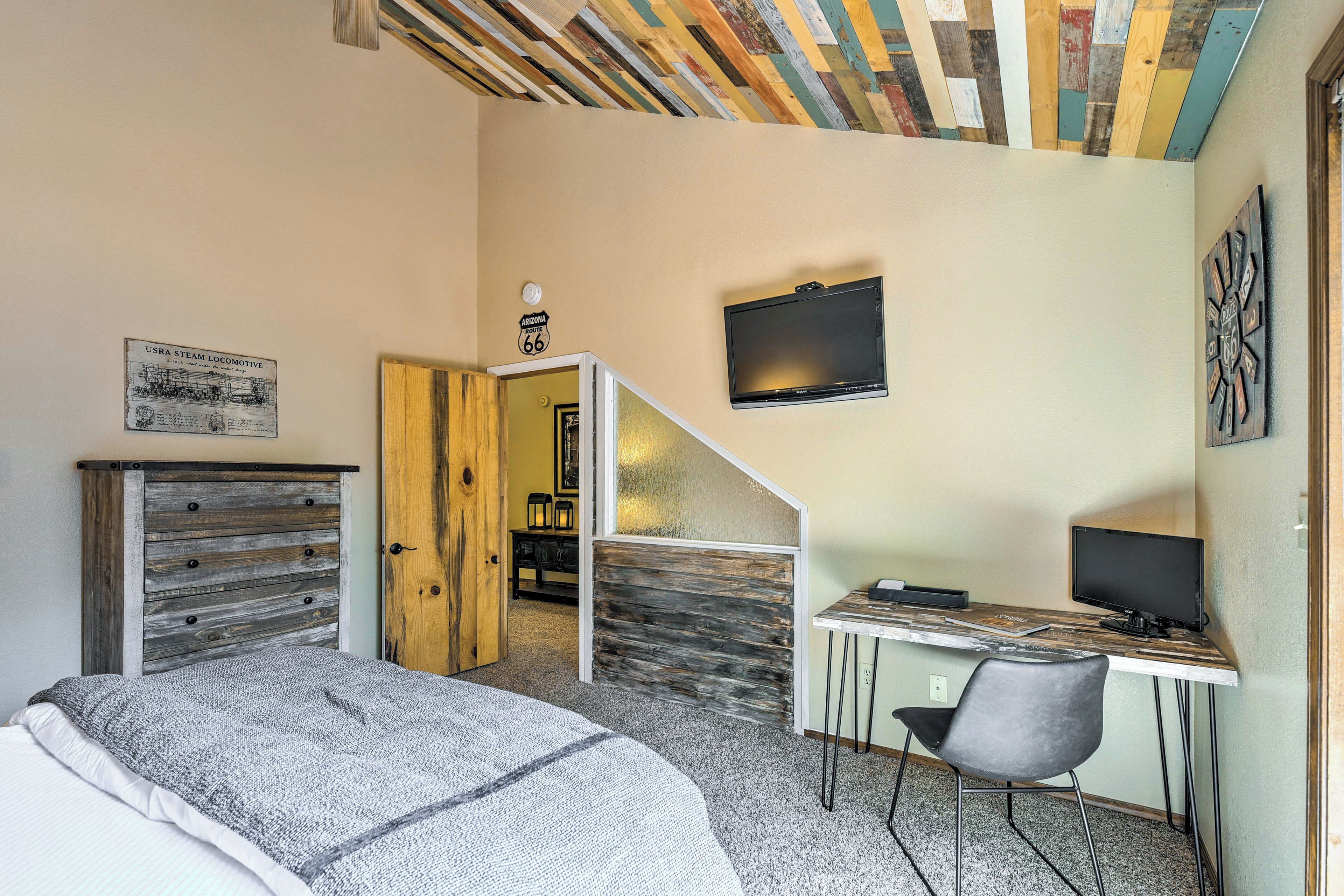 Ein Schlafzimmer mit einem Bett, einem Schreibtisch mit Computer, einem Sessel, einem Fernseher und einer Tür zu einem anderen Raum.