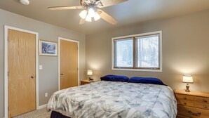 Ein Schlafzimmer mit einem Bett, einem Deckenventilator, zwei Fenstern mit Jalousien, einem Nachttisch mit Lampe und einer Holzkommode.