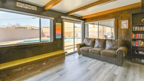 A cozy living area with a leather sofa, a bookshelf, and a large window offering a view of a swimming pool. - Location! Home w/ Pool 1 Min to Mcculloch Blvd (Lake Havasu City)