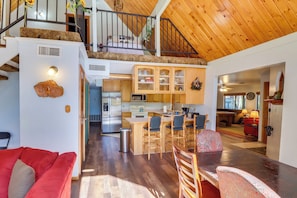 A spacious living area with a red sofa, wooden dining table, and a kitchen with wooden cabinets and a central island. - Relax Fireside: Rustic Gem 2 Mi to Rainbow Lake! (Lakeside)
