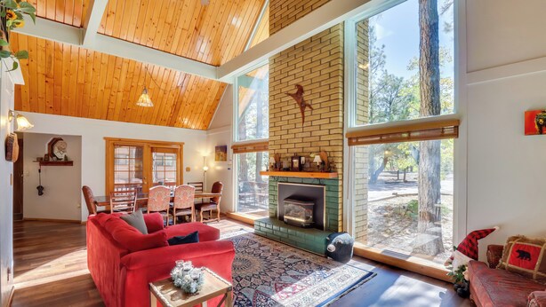 A spacious living room with a red sofa, a fireplace, and large windows offering a view of the outdoors. - Relax Fireside: Rustic Gem 2 Mi to Rainbow Lake! (Lakeside)