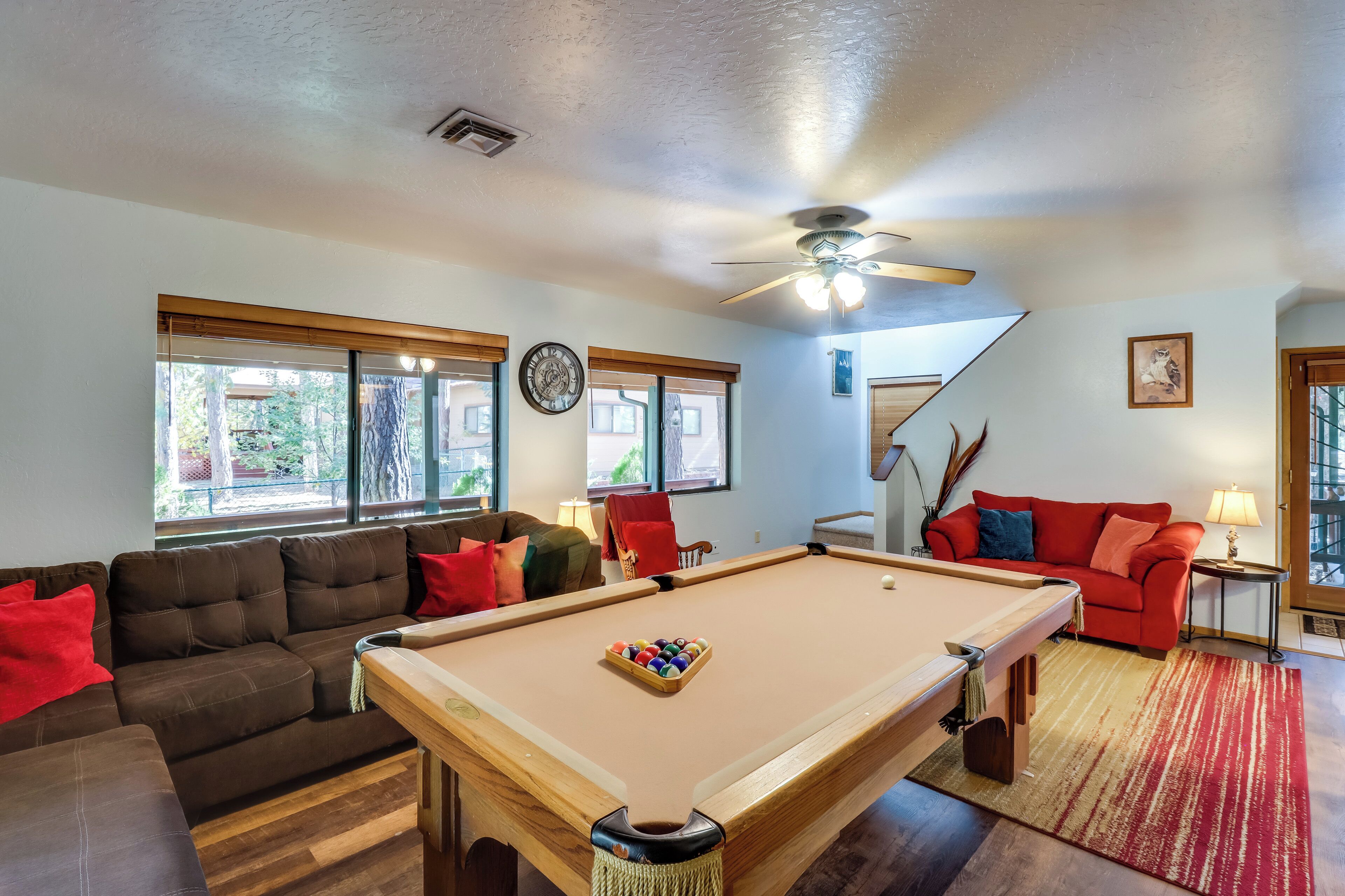 A pool table in a living room with a sofa and a red couch, large windows, and a ceiling fan.
