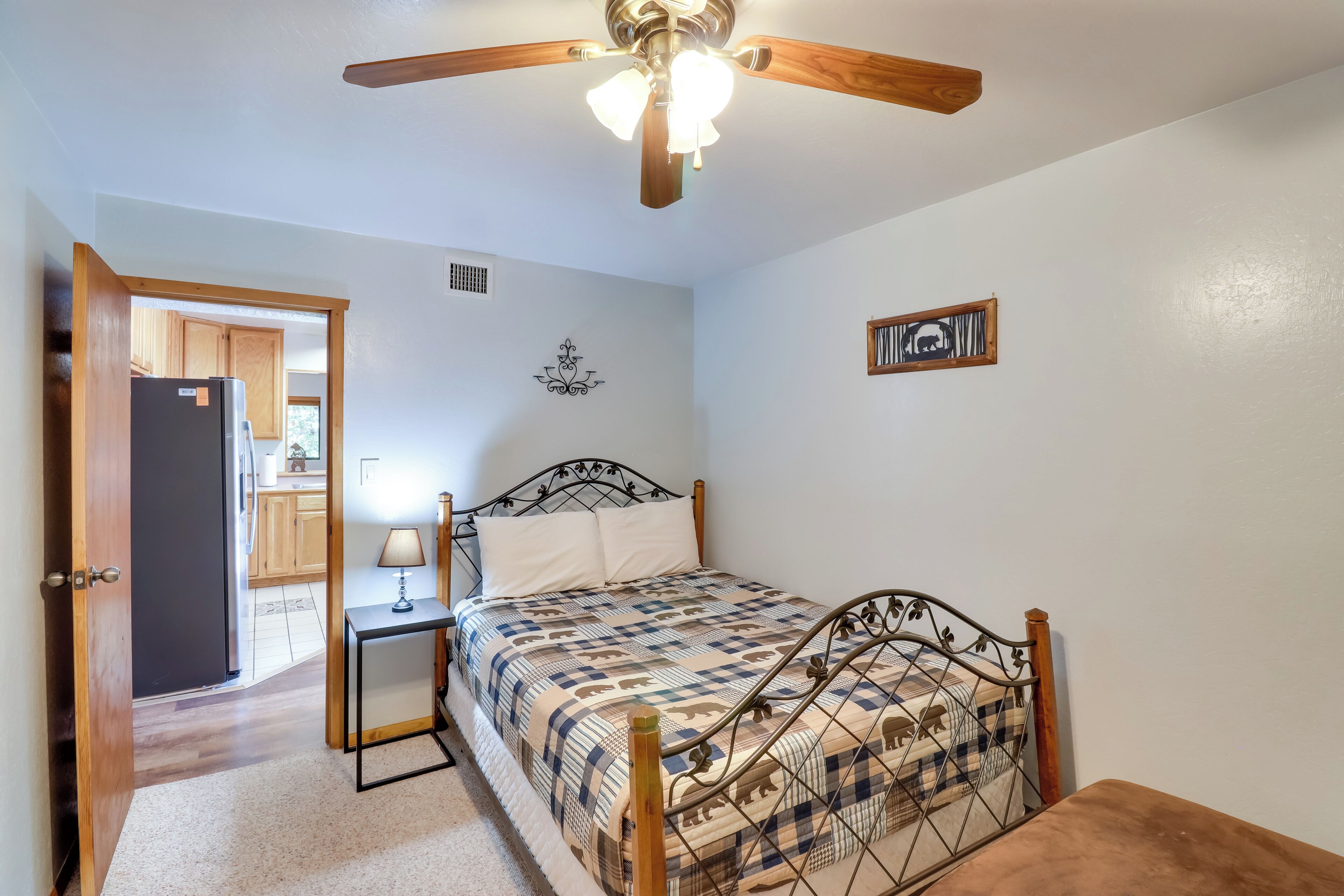 A bedroom with a bed, a nightstand, a ceiling fan, and a door leading to a kitchen.