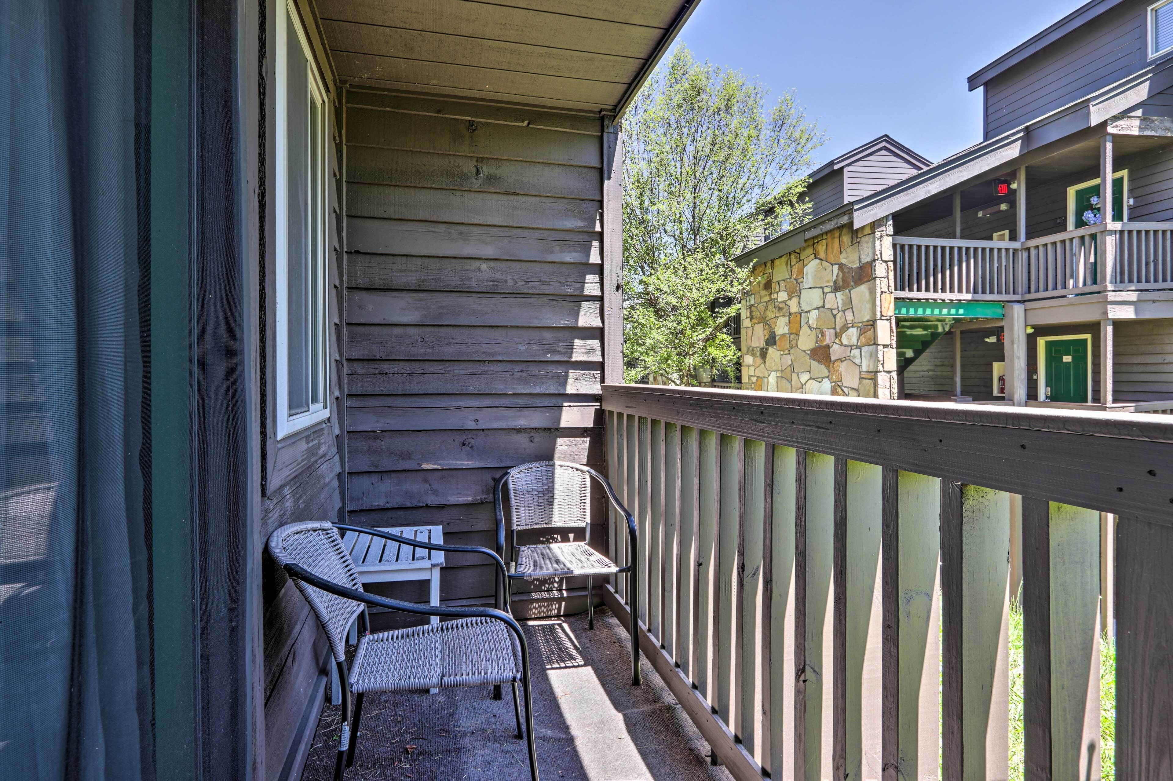 Een houten balkon met twee rieten stoelen en een tafeltje, met uitzicht op een stenen gebouw met een groene deur en een balkon.